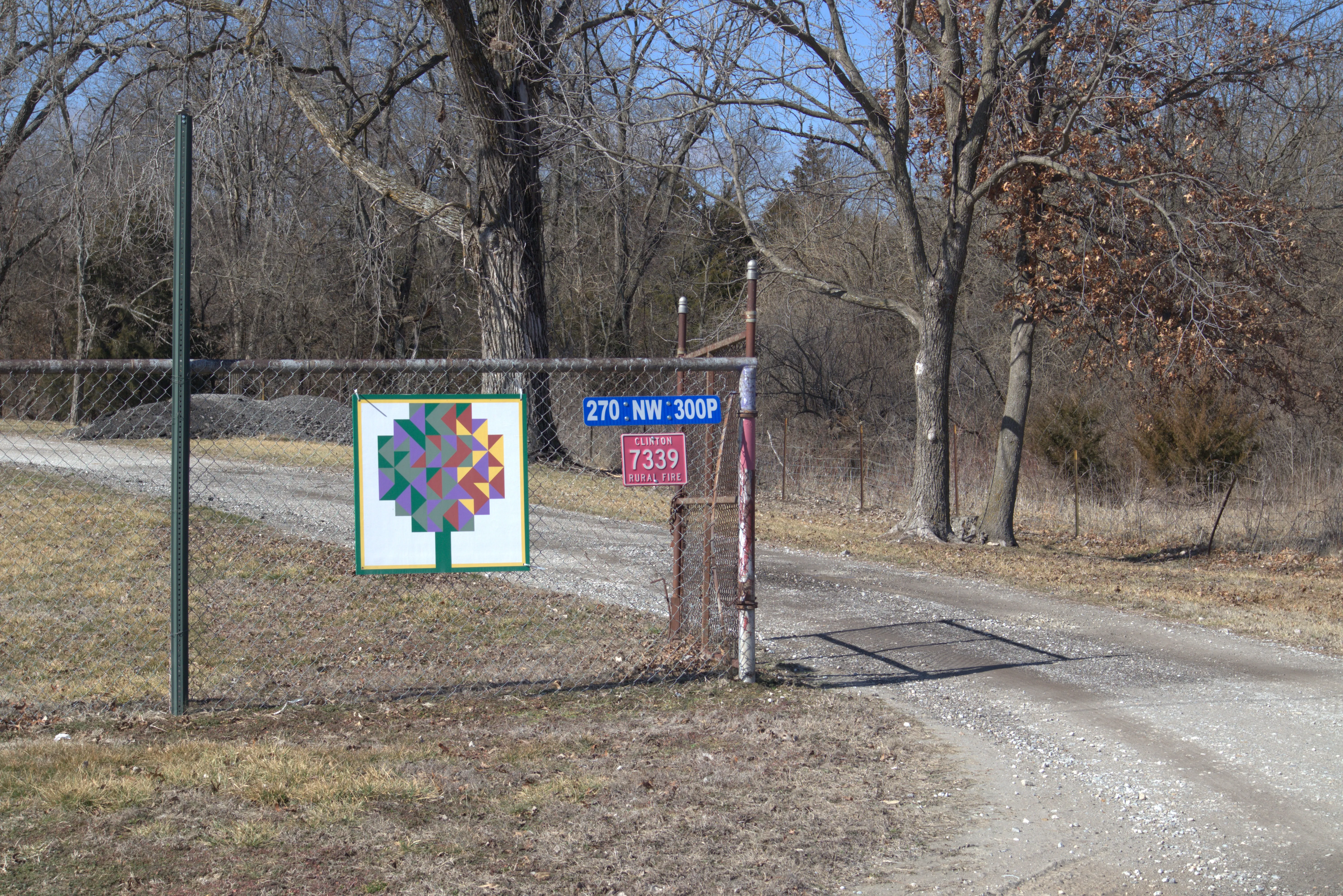 Retreat entrance gate with a colorful quilt barn block sign and address marker along a tree-lined gravel drive